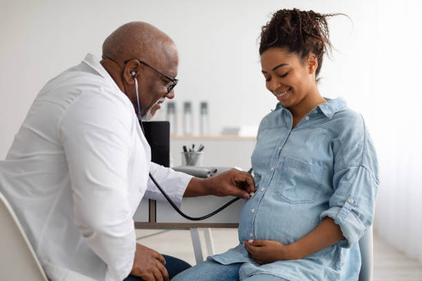 Smiling black pregnant woman visiting her obstetrician doctor in maternity clinic, mature male gynecologists examining her belly with stethoscope, doing medical check-up