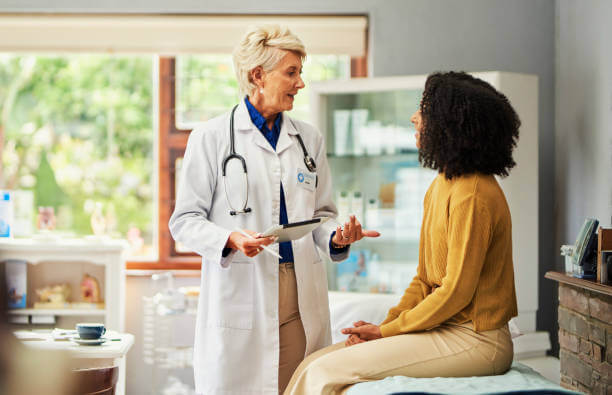 Female doctor holding digital tablet, consulting patient sitting on bed in clinic