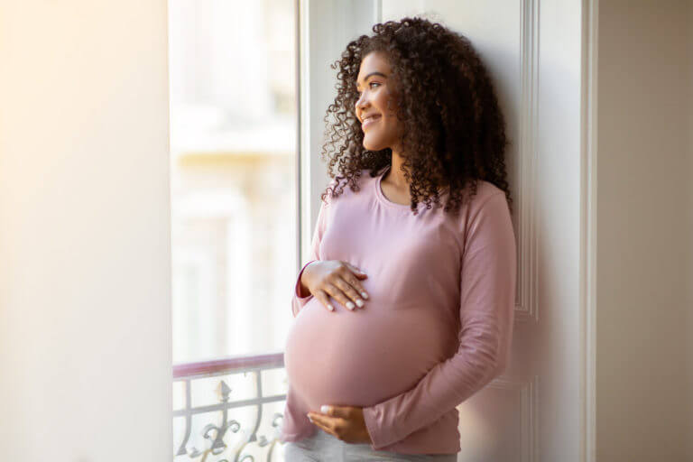 pregnant woman gently cradling belly while standing by window